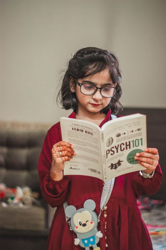 Home A young girl with eyeglasses and henna tattoos reading a book indoors.